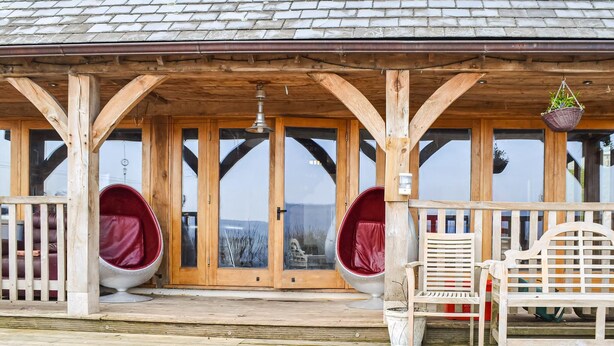 Un bâtiment en bois doté de grandes portes vitrées, de deux fauteuils rouges rembourrés et d’un banc blanc.