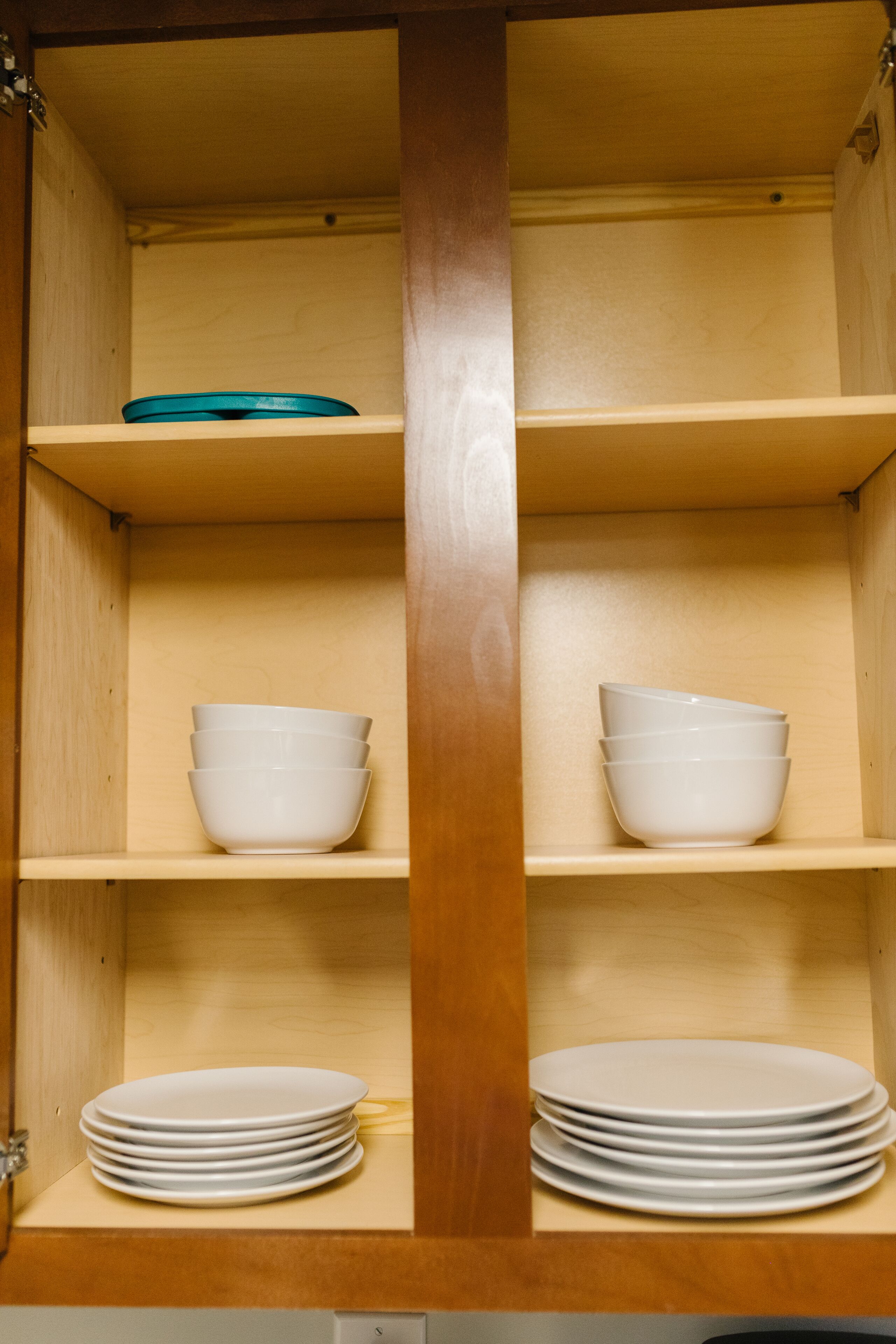 Un meuble en bois doté de trois étagères ; l’étagère supérieure contient un bol bleu, l’étagère du milieu abrite trois bols blancs, et l’étagère inférieure contient une pile d’assiettes blanches.