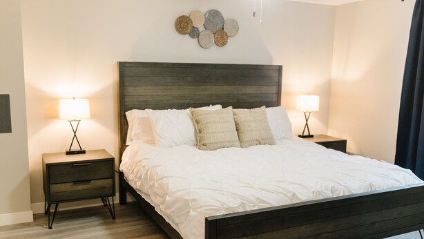 A neatly made bed with white linens, two bedside tables with lamps, and a decorative wall piece above the bed. - COZY BASEMENT APARTMENT - PRIVATE ENTRANCE - NEAR PARK (SALT LAKE CITY)