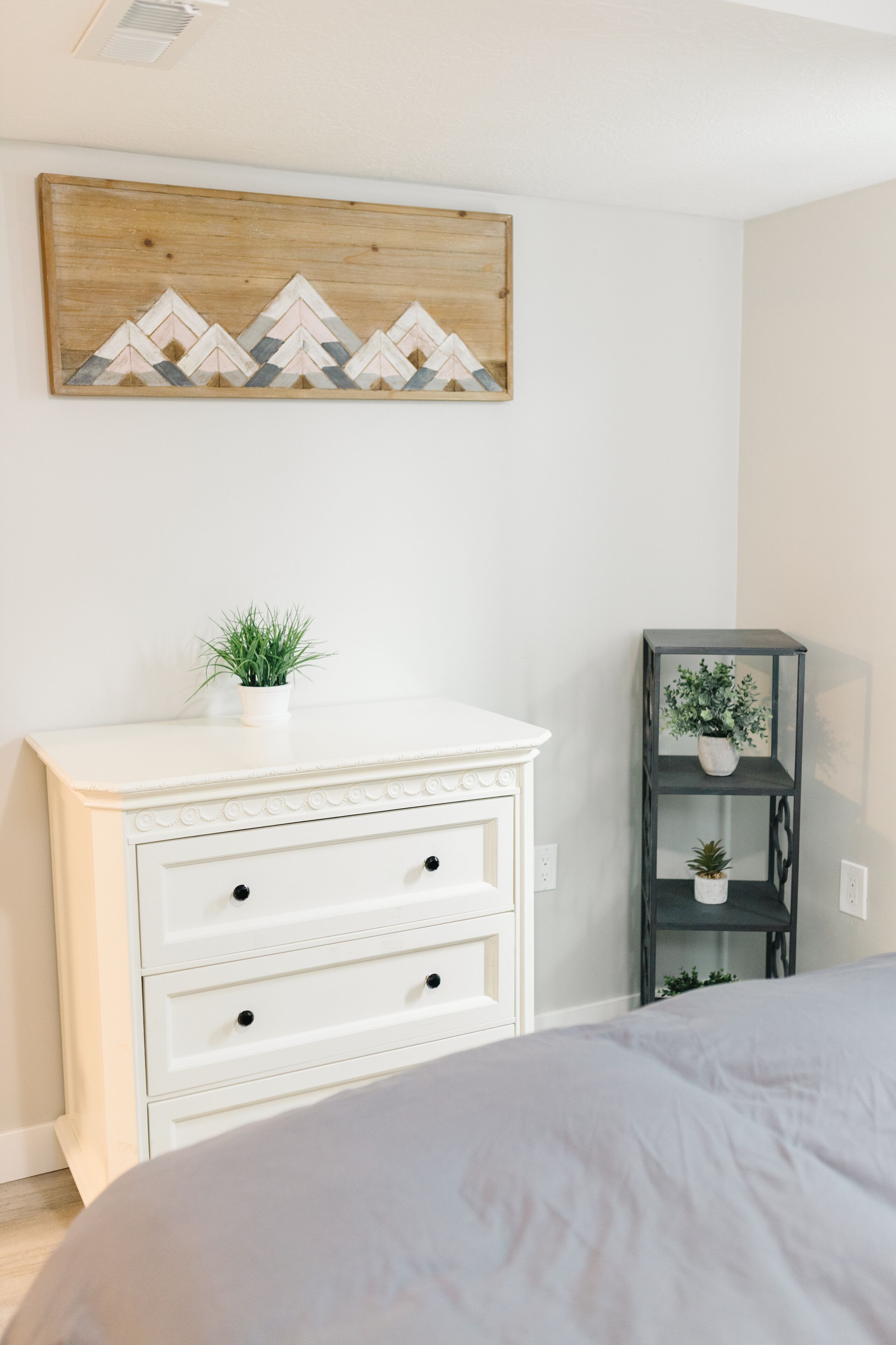 Un dormitorio con un aparador blanco, una cama, un pequeño estante con plantas y una obra de arte colgada en la pared.