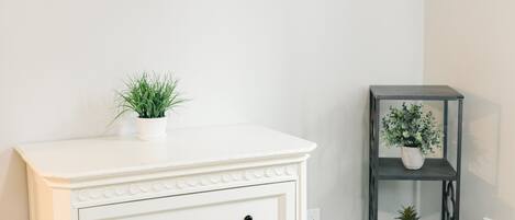 Un dormitorio con un aparador blanco, una cama, un pequeño estante con plantas y una obra de arte colgada en la pared.