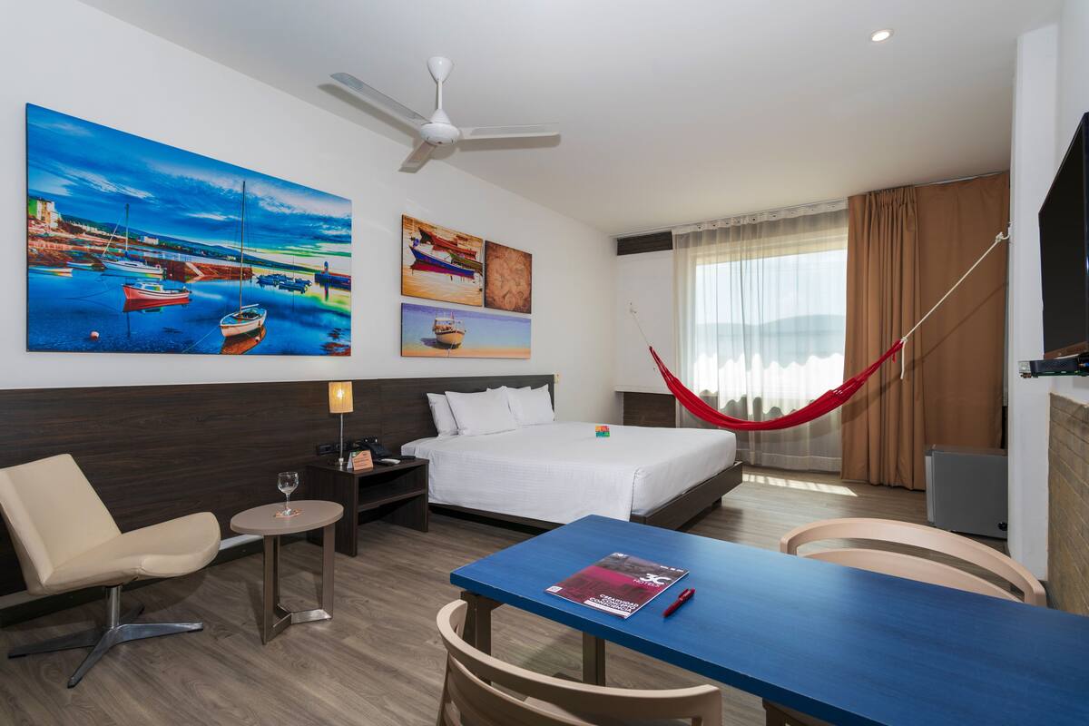 Une chambre d’hôtel avec un lit, un bureau, une chaise, une petite table, un hamac et des œuvres d’art accrochées aux murs.