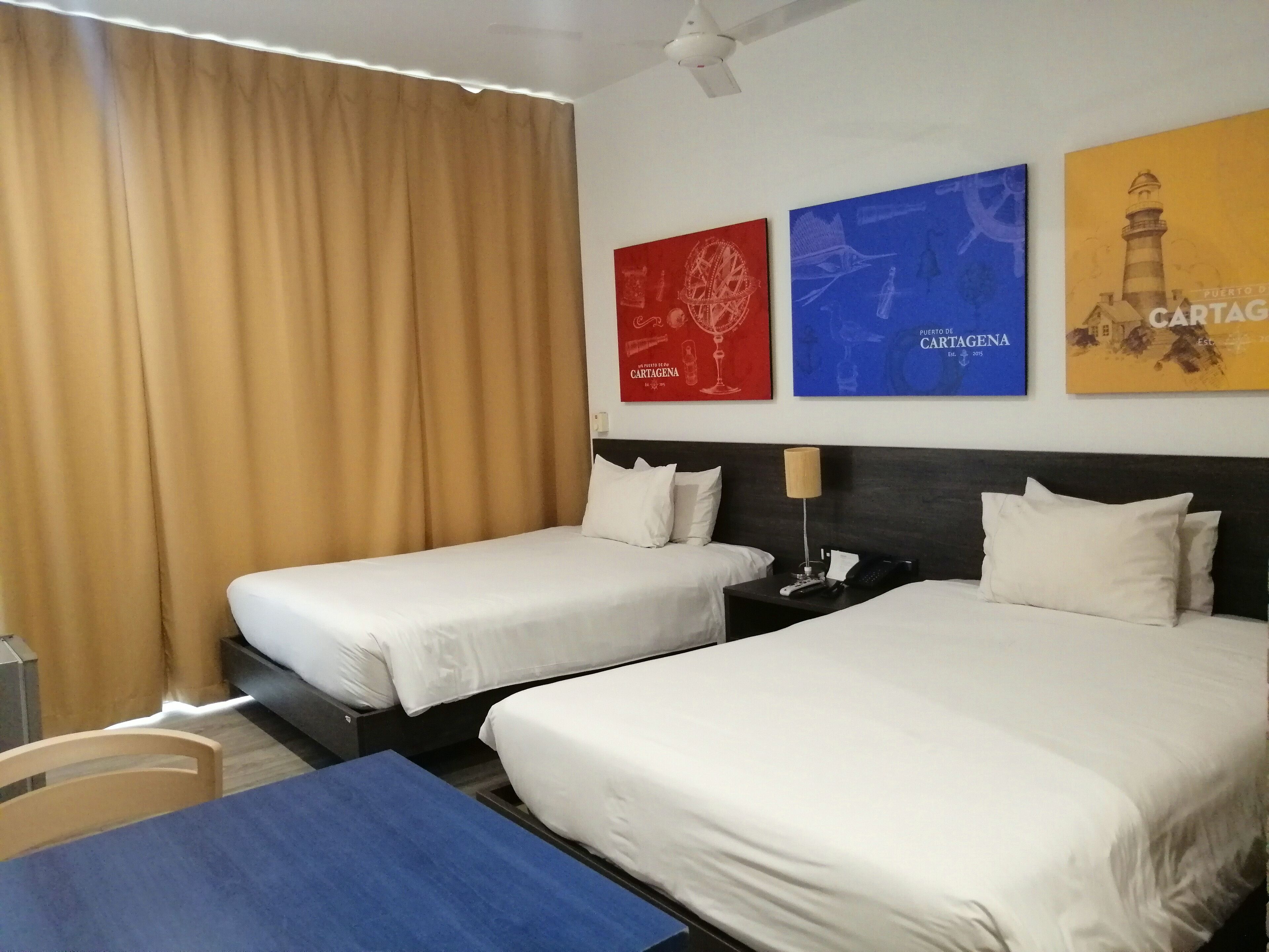 Habitación de hotel con dos camas, un escritorio y una silla. Hay tres carteles en la pared, uno rojo, uno azul y uno amarillo.