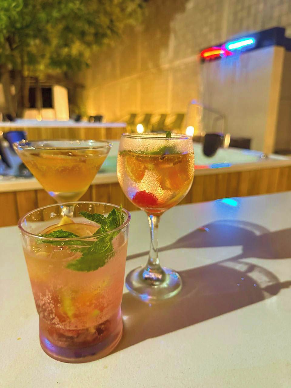 A bar area with three drinks on a table, a bar counter, and a wall with lights in the background.