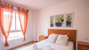 Ein ordentlich bezogenes Bett mit Kissen und aufgerollten Handtüchern, ein Holz-Kopfteil und ein Fenster mit orangefarbenen Vorhängen. Über dem Bett hängen drei gerahmte Bilder an der Wand.