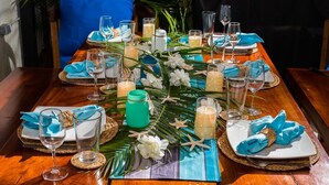 Een eettafel gedekt met blauwe servetten, glazen, kaarsen en tropische decoraties.