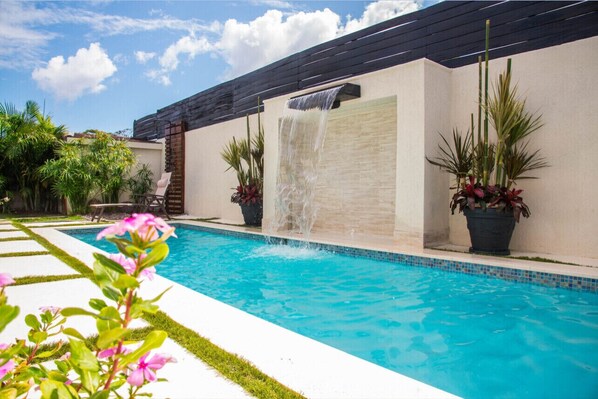 A swimming pool with a waterfall feature, surrounded by potted plants and a clear blue sky. - Modern Villa with private backyard pool (Drax hall)
