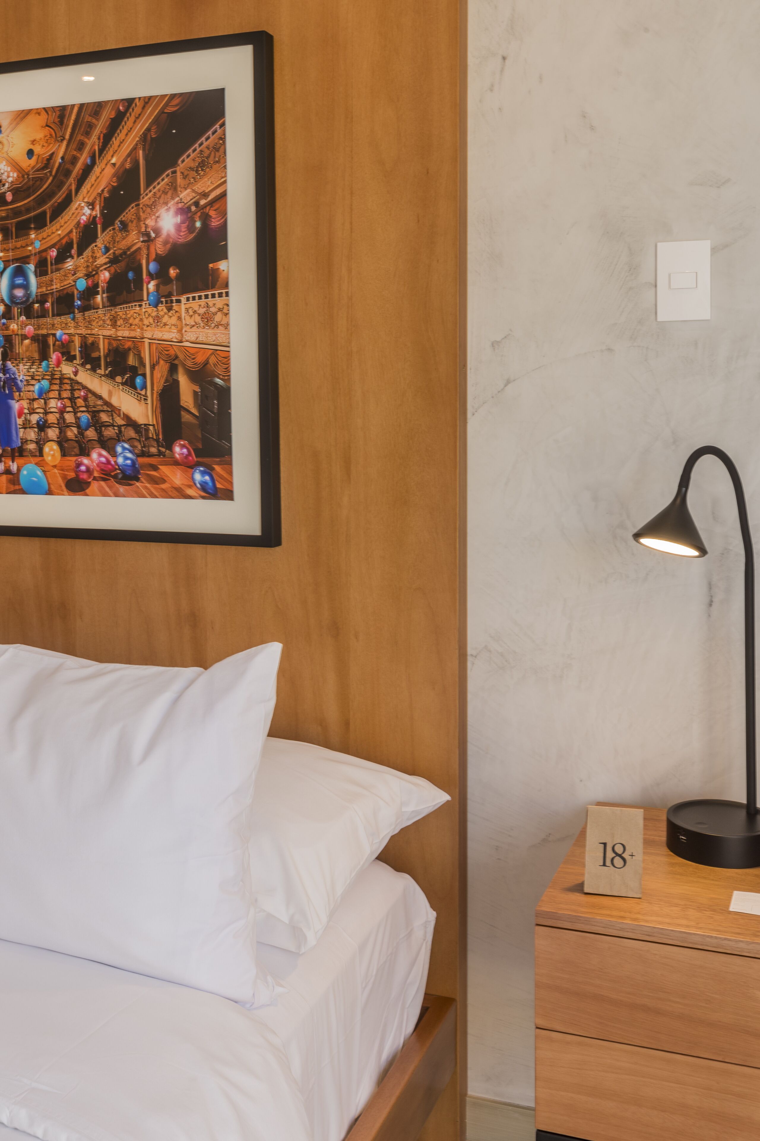 Ein Hotelzimmer mit Bett, Nachttisch mit Lampe, einem Bilderrahmen mit Theaterabbildung und einer Holzwandverkleidung.