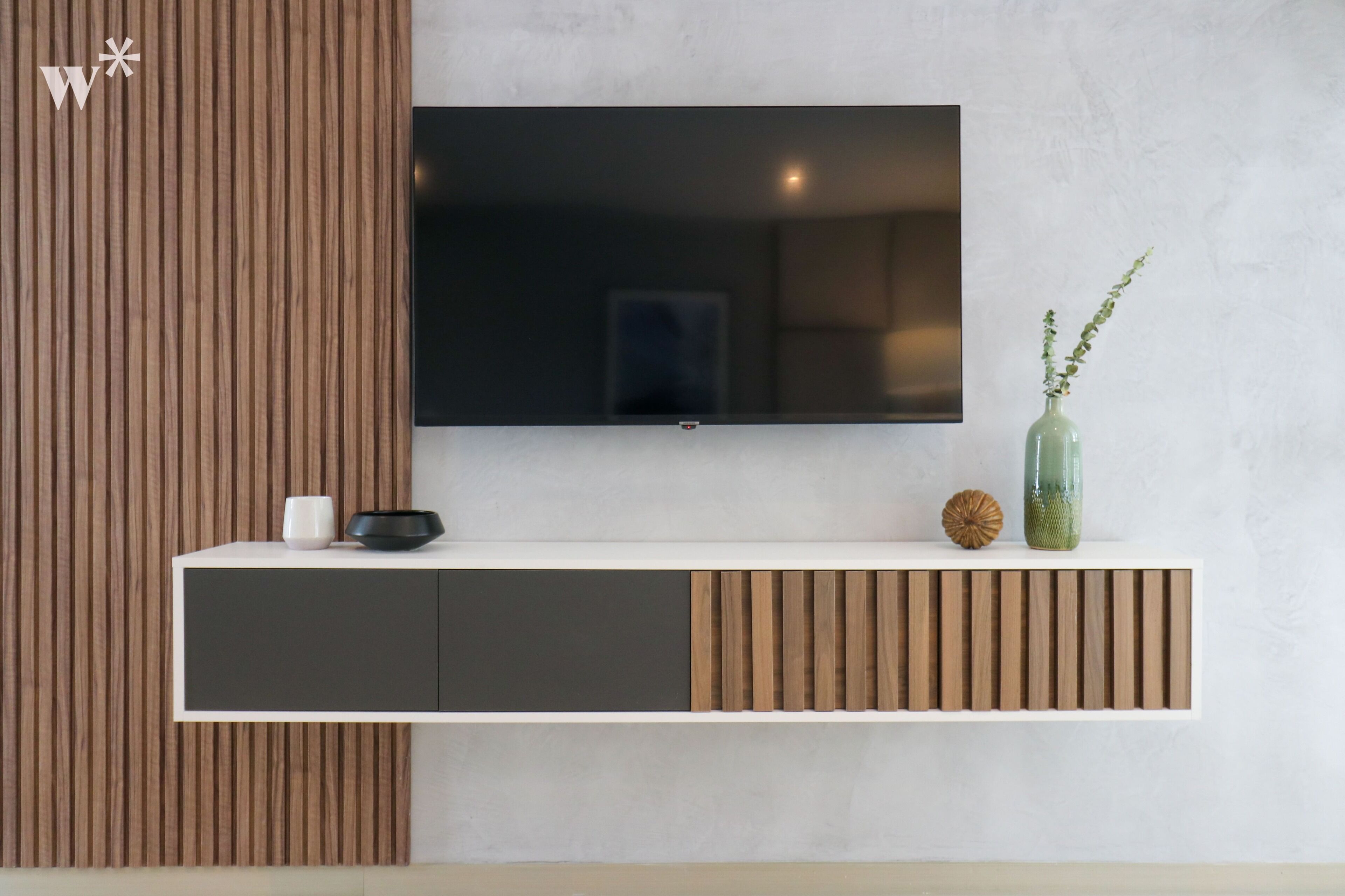 A modern living room with a flat-screen TV mounted on a white wall, a wooden accent panel, a white console table with a black bowl and a green vase with a plant, and a decorative sphere.