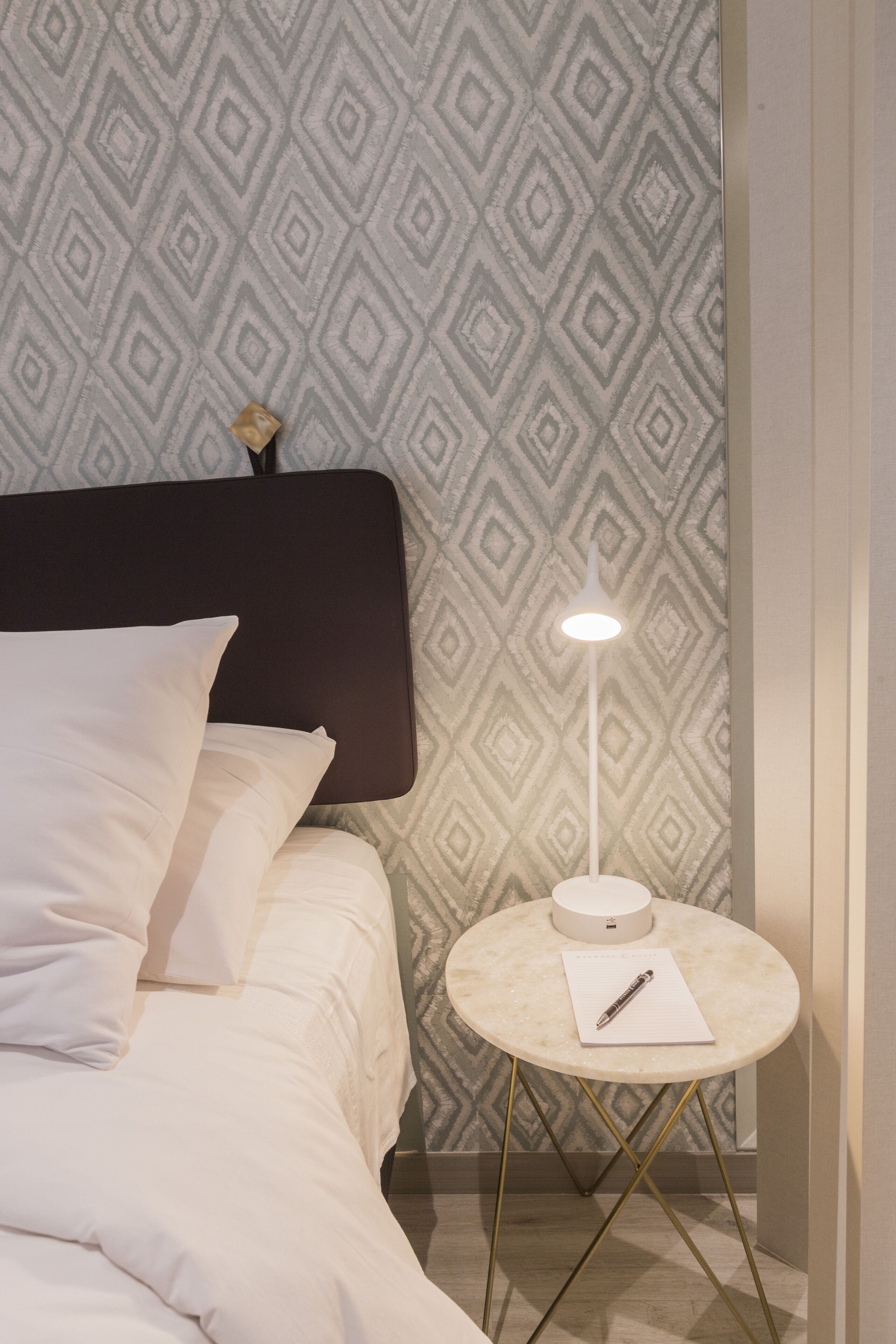 A neatly made bed with white linens, a round side table with a white lamp, and a patterned wallpaper in the background.