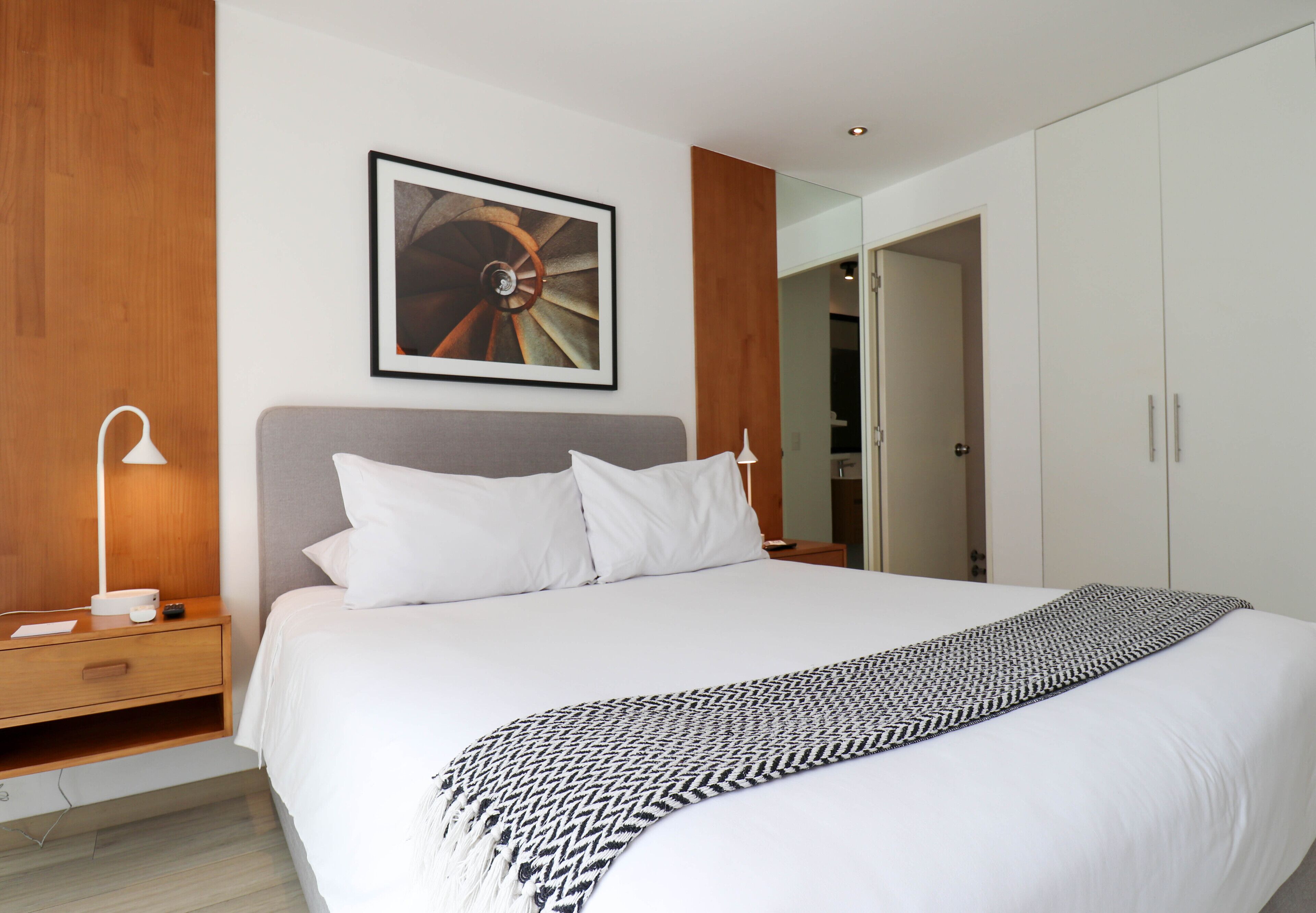 A modern hotel room with a large bed, a bedside table with a lamp, a framed picture on the wall, and a wardrobe.