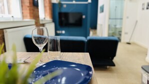 A dining table with a blue plate and two wine glasses, a brick wall, a blue sofa, and a television in the background. - Lovely Studio Apartment in the Heart of Nottingham (Nottingham)