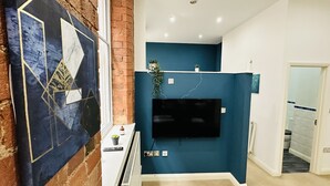 A modern living room with a large TV mounted on a blue accent wall, a white radiator, and a brick wall with a geometric painting. - Lovely Studio Apartment in the Heart of Nottingham (Nottingham)