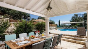 A covered outdoor dining area with a pool in the background. - Villa Perla by Villa Plus (Kassiopi)