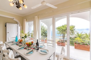 A dining area with a table set for four, large windows with a view of the sea, and a ceiling fan.