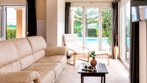 Ett modernt vardagsrum med en beige lädersoffa, en trästol och ett litet bord med en vas och ett par solglasögon. Stora fönster ger utsikt över en swimmingpool och grönområden.