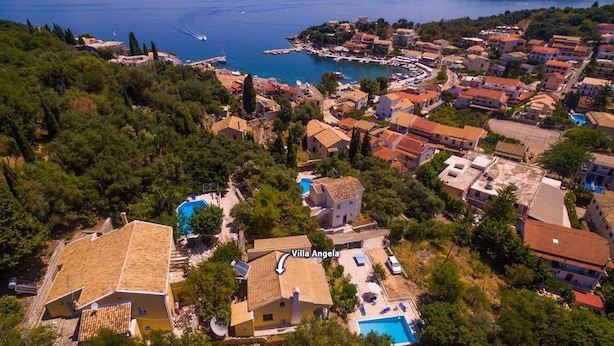 Ovenudblik over Villa Angela med en swimmingpool, omgivet af bygninger i middelhavsstil og en kystlinje i baggrunden.