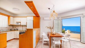 Eine moderne Küche mit weißen Schränken und einer Holztischinsel, ein Essbereich mit weißen Stühlen und einem Holztisch sowie ein Balkon mit Meerblick.