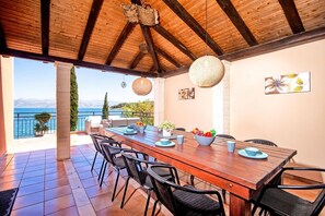 A wooden dining table with chairs, a view of the sea, and a balcony with a railing. - Villa Annie by Villa Plus (Imerolia)