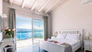 Ein Schlafzimmer mit Bett, Nachttischen, einem Schreibtisch und einem Sessel. Ein großes Fenster bietet Blick auf ein Gewässer und Berge.
