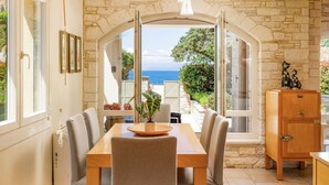 Zona pranzo con parete in pietra, tavolo in legno e sedie. Vista sul mare attraverso una finestra ad arco.