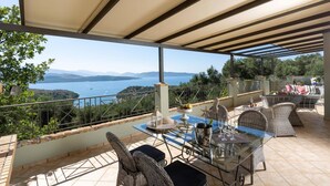 An outdoor patio with a glass table, wicker chairs, and a view of a lake and mountains. - Villa Firefly by Villa Plus (Άγιος Στέφανος)