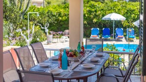 An outdoor dining area with a pool view, tables set with plates and glasses, and a variety of fruits and bottles. - Villa Foula by Villa Plus (Άγιος Στέφανος)