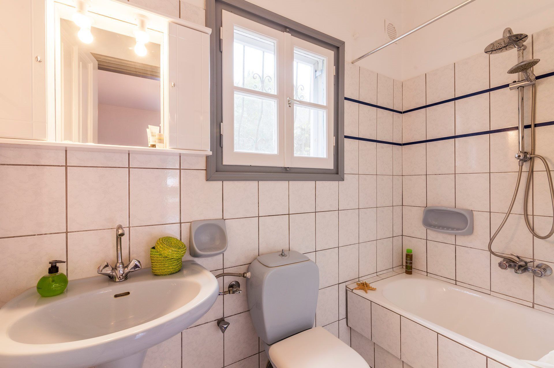 Een badkamer met een wastafel, toilet en bad. Er is een raam boven de wastafel en een douchekop boven het bad.