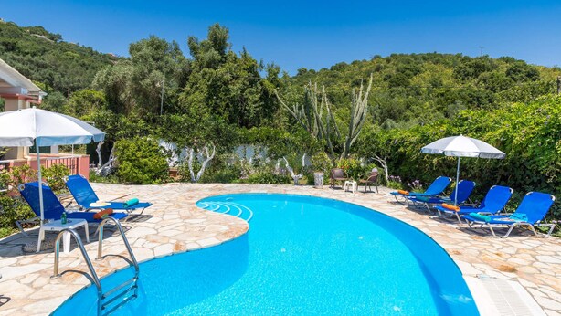 A swimming pool with blue water, surrounded by stone tiles. There are blue lounge chairs and white umbrellas. - Villa Foula by Villa Plus (Άγιος Στέφανος)