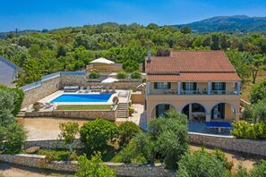 A two-story house with a swimming pool, stone walls, and a view of the surrounding greenery and hills.