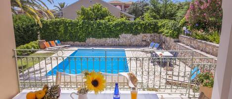 A balcony with a view of a swimming pool, a table with fruits, a pitcher, and glasses.