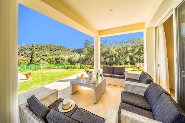 Un espace extérieur aménagé, abrité et doté de mobilier en osier et d’une table en verre, offrant une vue sur un jardin paysager et des collines.