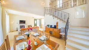 A spacious dining area with a high ceiling, a staircase with ornate railings, and a view of the outdoors through large windows. - Villa Gina by Villa Plus (Ionian Islands)