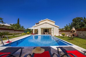 Vue d’une maison avec façade en pierre, un patio couvert et un espace barbecue, le tout vu depuis la piscine.