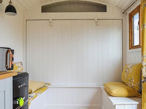 Une pièce avec un banc en bois blanc, des coussins jaunes, un appareil noir portant l’inscription « HAUS » et une fenêtre avec des rideaux jaunes.