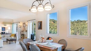 A dining area with wicker chairs, a wooden table, and a view of a forest through large windows. - Villa Kalliopi by Villa Plus (Kokkini)