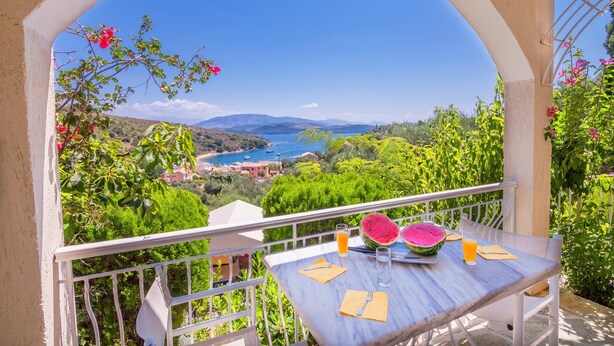 A balcony with a table set for two, overlooking a coastal landscape with houses and a clear blue sea. - Villa Kerkyra by Villa Plus (Άγιος Στέφανος)