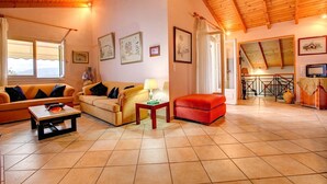 A spacious living room with a high wooden ceiling, tiled floor, and a cozy seating area with a sofa and armchairs. - Villa Kerkyra by Villa Plus (Άγιος Στέφανος)