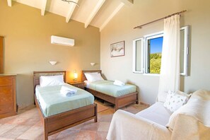 A bedroom with two wooden beds, a wooden dresser, a window with a view of greenery, and an air conditioning unit. - Villa Kokokilas by Villa Plus (Ionian Islands)