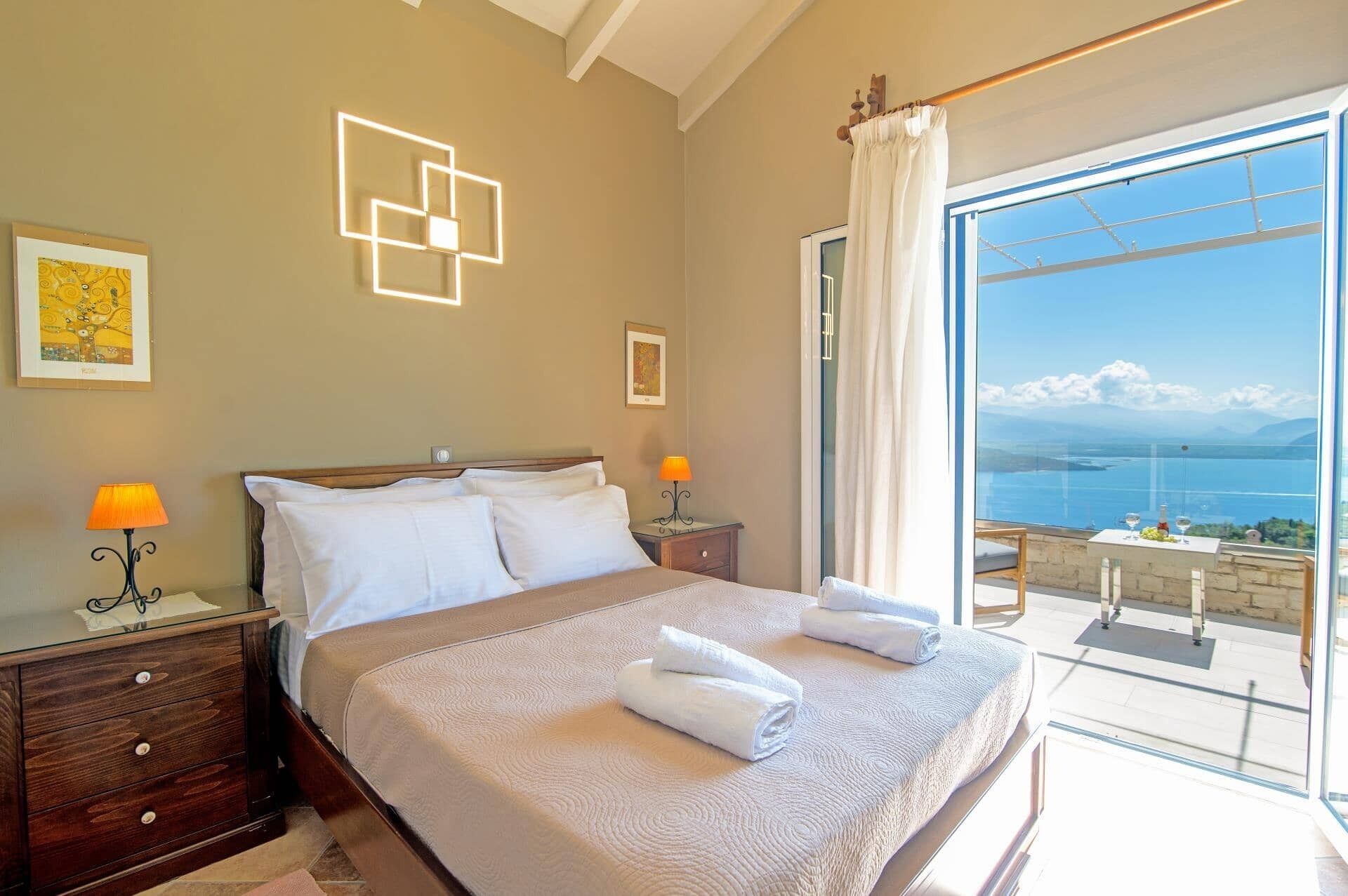 A bedroom with a large bed, two bedside tables with lamps, a view of the sea through a balcony, and artwork on the walls.