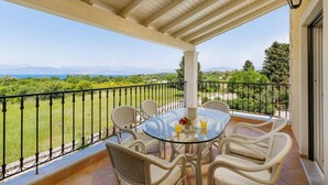 A balcony with a glass table and wicker chairs, overlooking a lush green landscape and distant mountains.