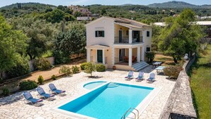 A two-story villa with a swimming pool, surrounded by greenery and hills.