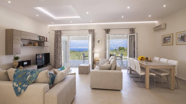 A modern living room with a sofa, a TV, and a dining area with a table and chairs. There are curtains and a view of the outdoors.