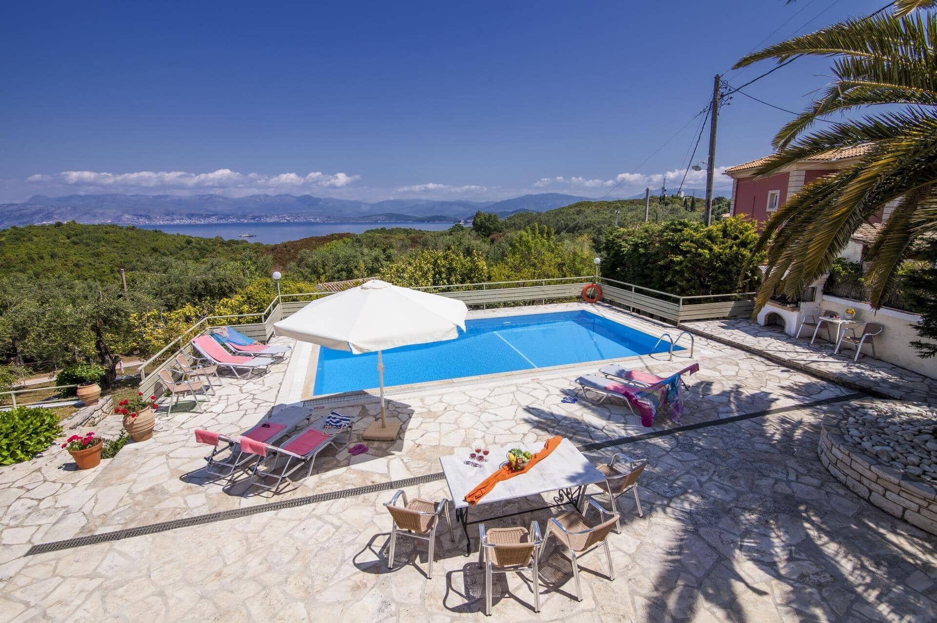 A pool area with lounge chairs, a table with drinks, and a view of the sea and mountains.