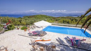 A pool area with lounge chairs, a table with a vase of flowers, and a view of the sea and mountains.