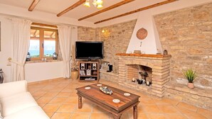 Uma sala de estar com lareira de pedra, vigas de madeira, televisão e vista pela janela.