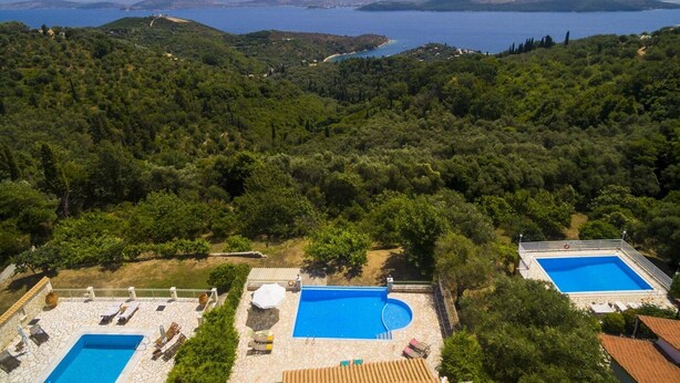 Et resort med to swimmingpools, omgivet af frodig vegetation og udsigt over et stort vandområde.