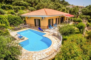 A villa with a swimming pool, surrounded by greenery and a red-roofed building.
