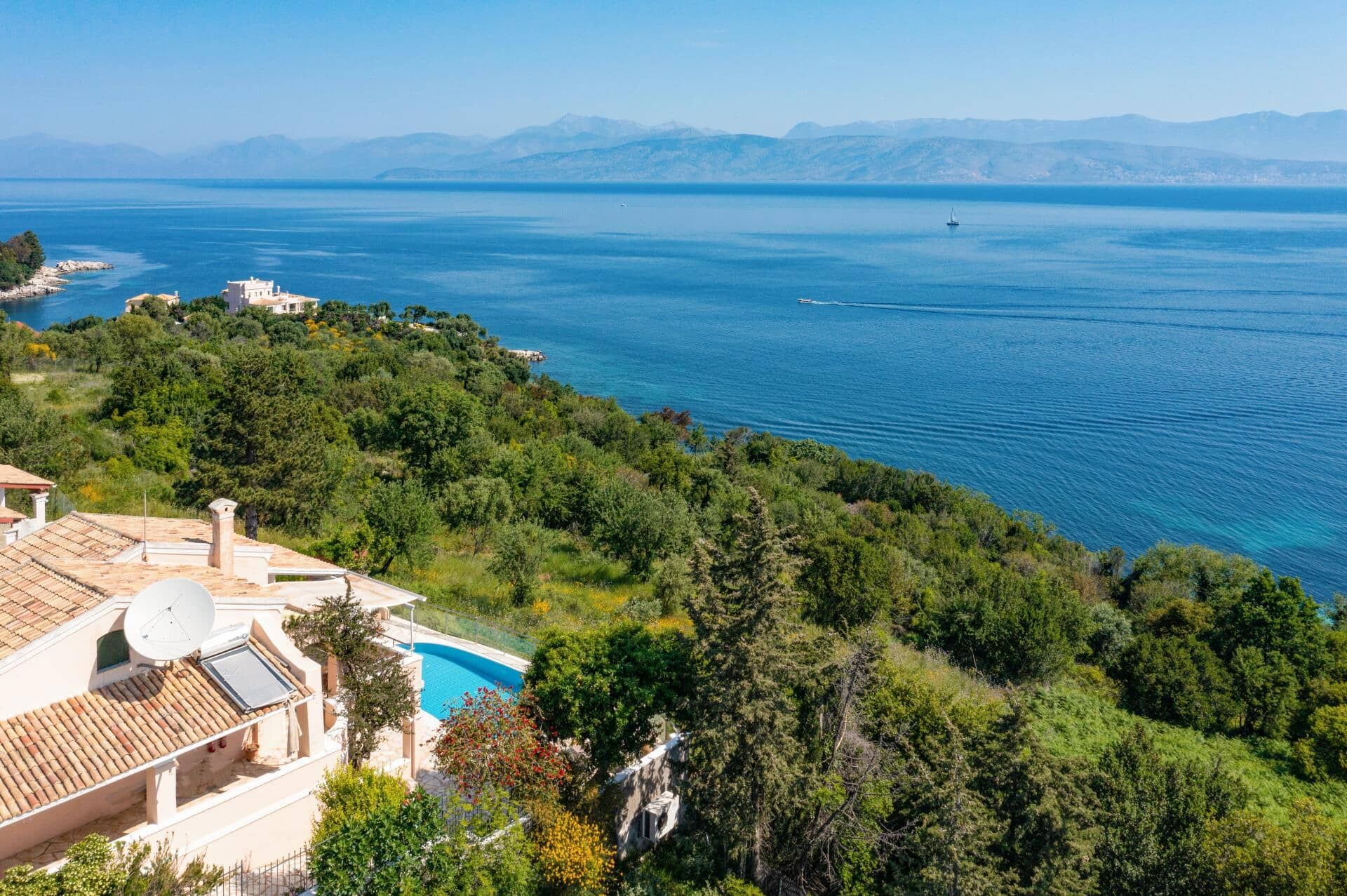 A coastal property with a swimming pool, lush greenery, and a view of the sea and distant mountains.