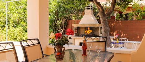 Eine Terrasse im Freien mit einem Glastisch, Stühlen, einer Vase mit Blumen, einer Flasche und einem Glas. Im Hintergrund steht ein Grill und ein Baum.
